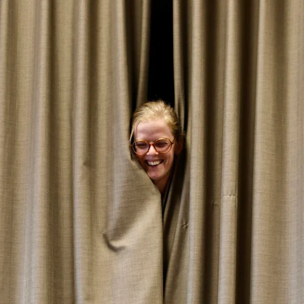 Beeld: Lachend gezicht van een vrouw met bril, dat komt piepen van achter een beige theatergordijn.